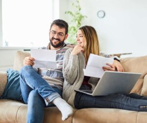 Couple at home on the couch looks at paperwork and laptop to consider the pros and cons of refinancing their home.