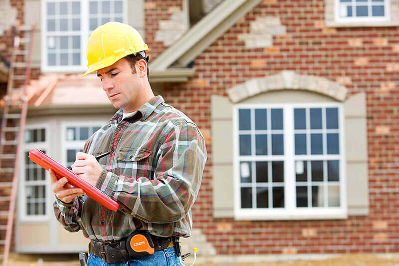 Home inspector in a hard hat reviewing a home inspection checklist in front of a brick house under evaluation