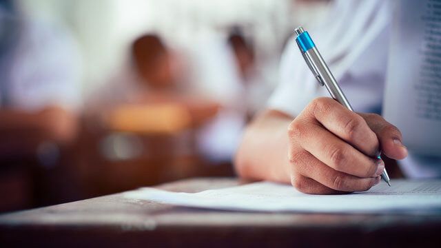 Close up of an arm and a hand of someone sitting at a desk in a classroom. They are holding a pen and are taking an exam.