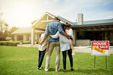 A family in the front yard of their new home. A for sale sign next to them with a sold sign places over it.