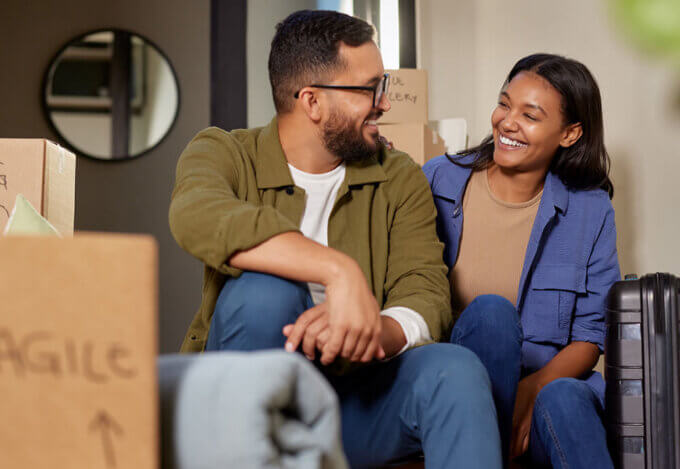 A smiling couple sitting among moving boxes and luggage, planning their next home purchase before selling their current one.