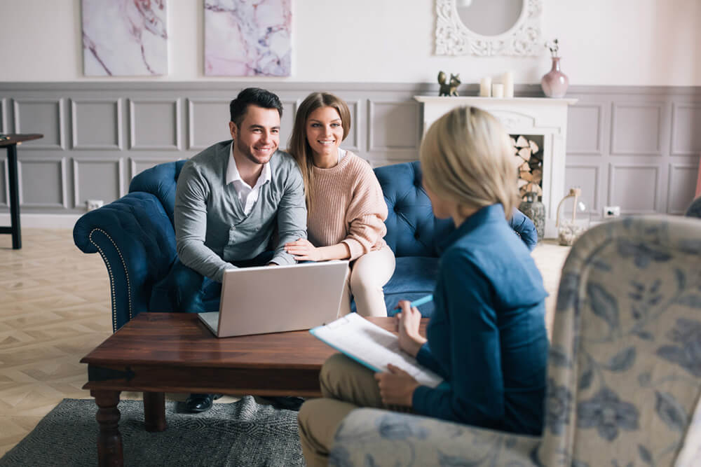 A couple meeting with a realtor. They are sitting across from the realtor on a couch looking at a laptop.