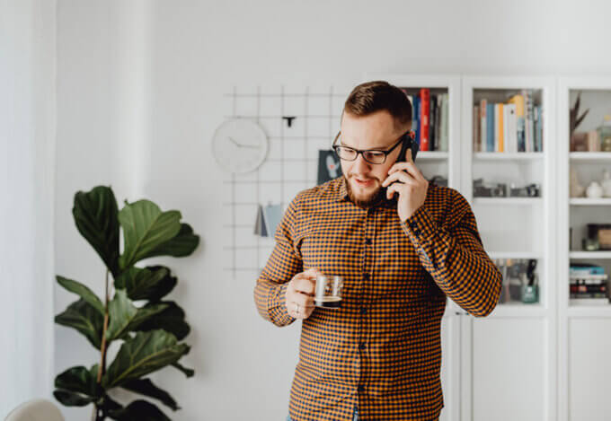 A man standing in his living room. On the phone while holding a small coffee cup.