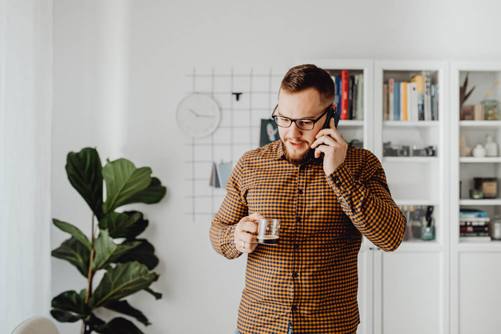 A man standing in his living room. On the phone while holding a small coffee cup.