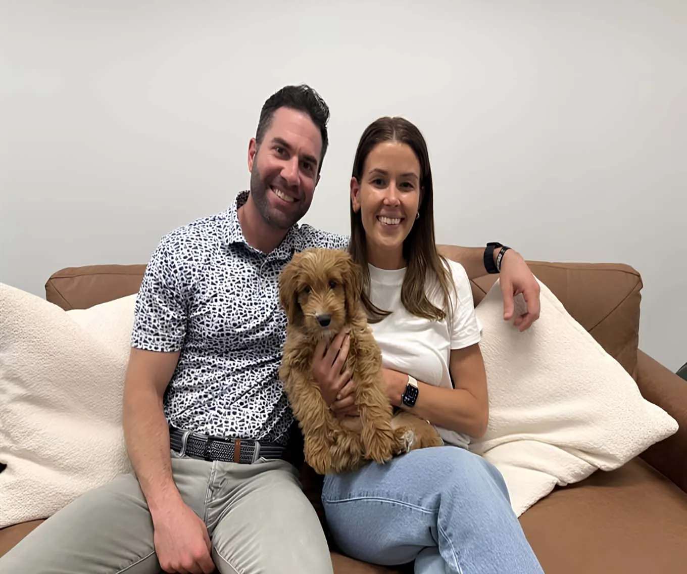 A couple sitting on a couch, holding a goldendoodle puppy.
