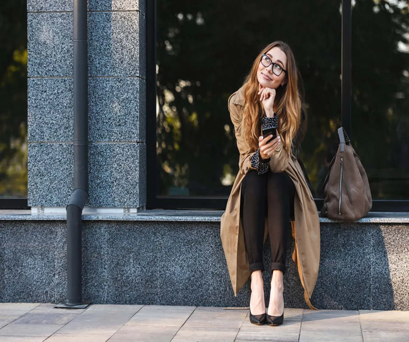 A woman sitting on a step of a building. She is holding her phone looking up and thinking.