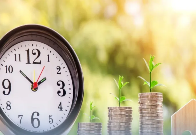A small clock next to three stacks of coins with a plant sprouting out of each.