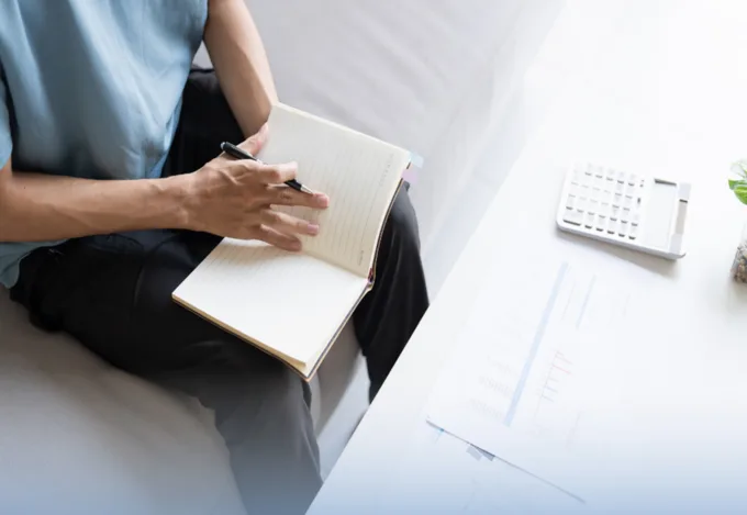 A potential homebuyer sitting on their couch, holding an open notebook. A calculator on the coffee table in front of them.