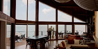 Couple standing at the entrance to a waterfront deck, viewed from inside a wood-paneled room with floor-to-ceiling windows