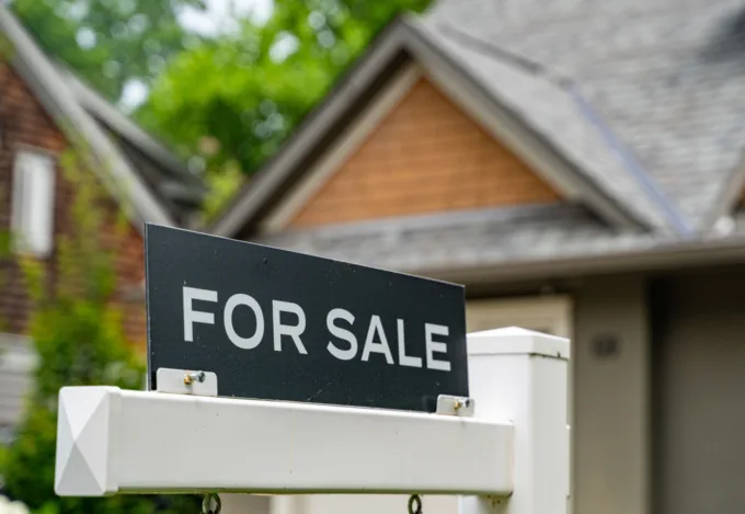 For sale sign displayed in front of a suburban house with a peaked roof, front porch and green trees in the background.