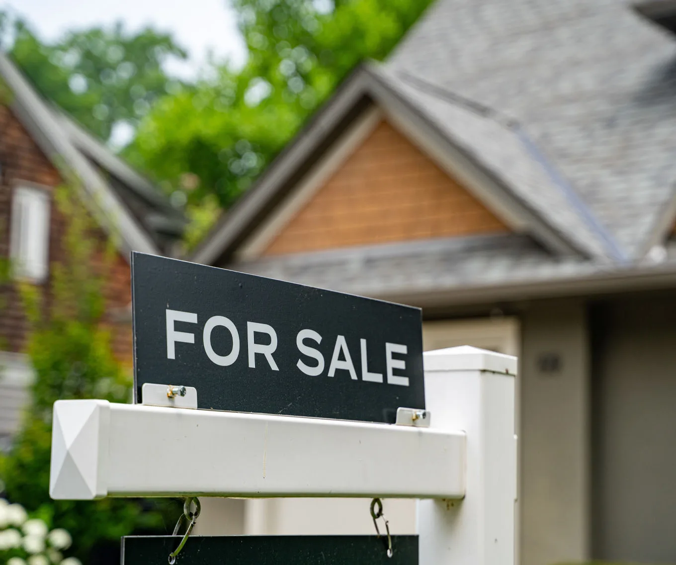 For sale sign displayed in front of a suburban house with a peaked roof, front porch and green trees in the background.