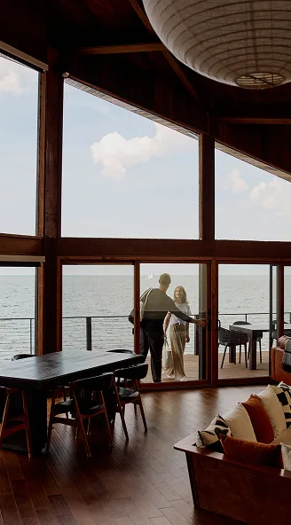 Couple standing at the entrance to a waterfront deck, viewed from inside a wood-paneled room with floor-to-ceiling windows