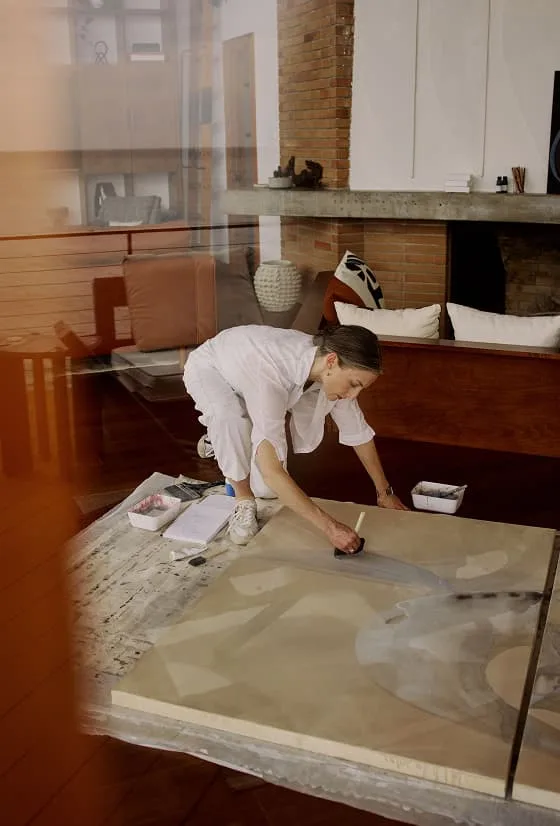 Woman working on a large artwork spread across a table in a living room.