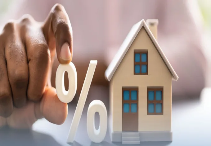 Hand holding a percent sign next to a small model house, representing mortgage interest rates