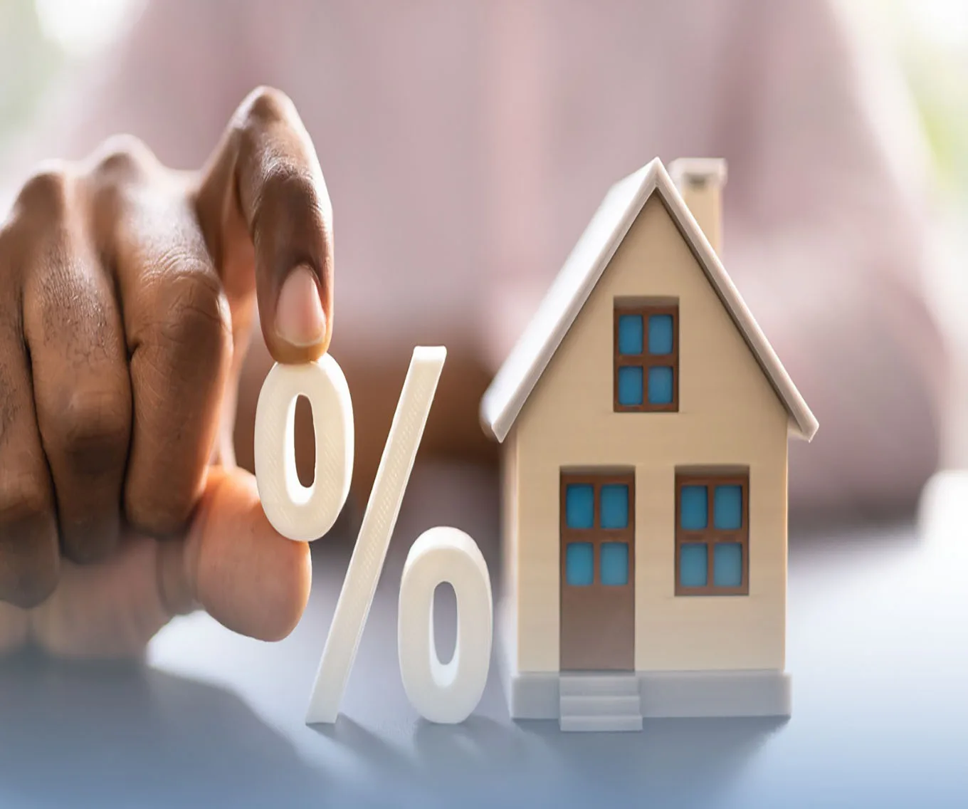 Hand holding a percent sign next to a small model house, representing mortgage interest rates