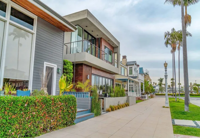 Modern beachside homes with balconies and lush landscaping along a palm-tree-lined sidewalk in a coastal neighborhood