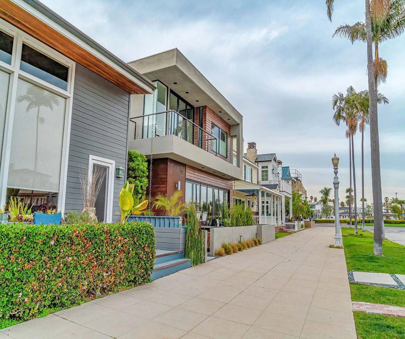 Modern beachside homes with balconies and lush landscaping along a palm-tree-lined sidewalk in a coastal neighborhood