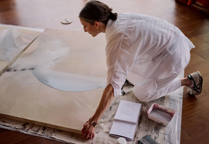 Artist kneeling on a studio floor, working on a large canvas painting with soft blue shapes, surrounded by a sketchbook and paint supplies