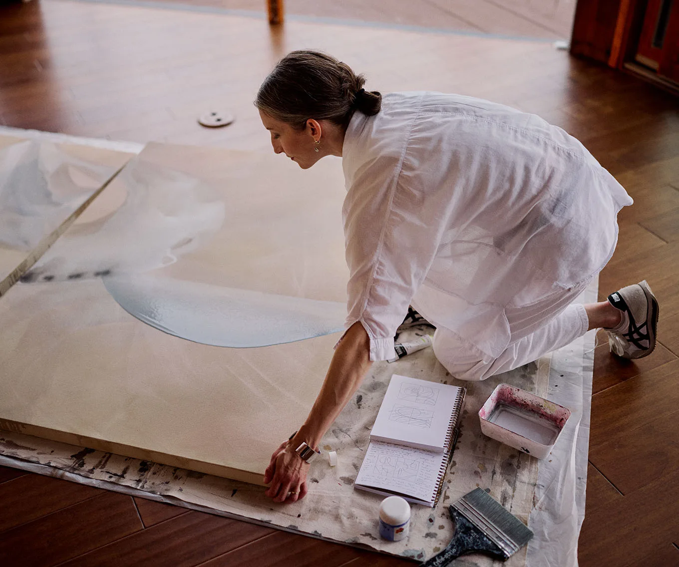 Artist kneeling on a studio floor, working on a large canvas painting with soft blue shapes, surrounded by a sketchbook and paint supplies
