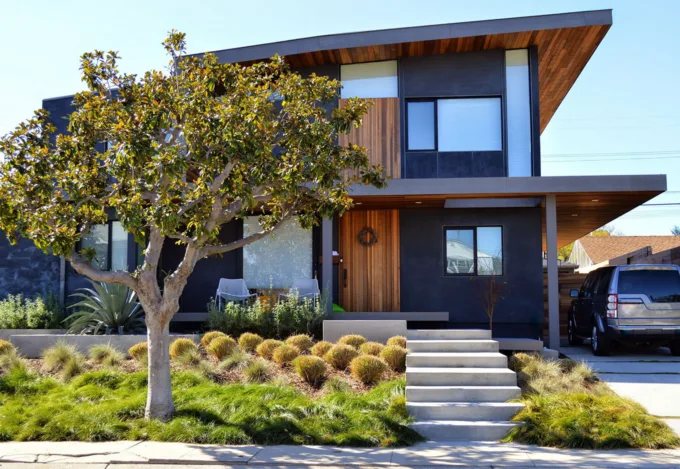 Modern two-story home with dark exterior, wood accents, flat roof overhang, concrete steps, and drought-tolerant landscaping