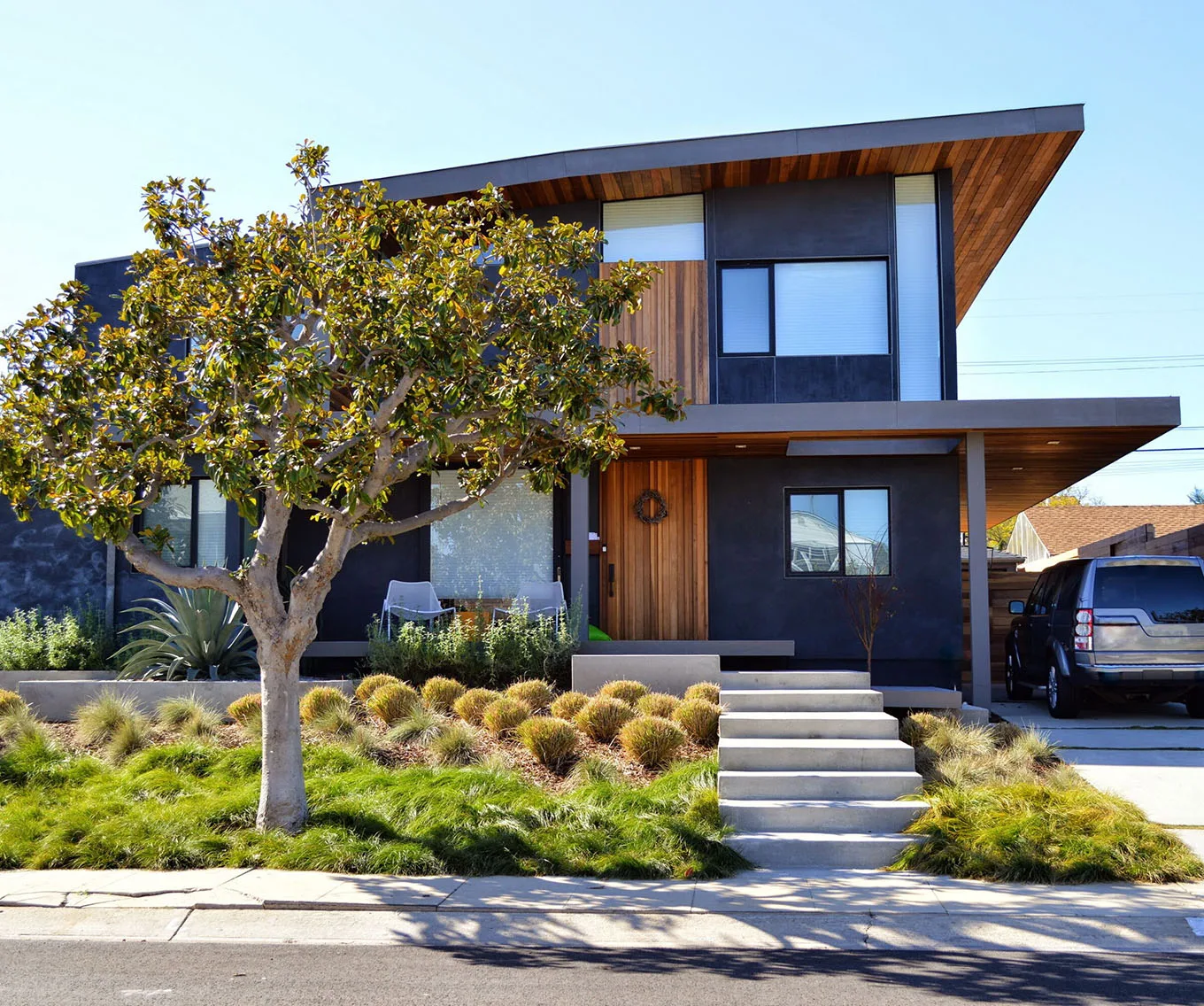 Modern two-story home with dark exterior, wood accents, flat roof overhang, concrete steps, and drought-tolerant landscaping
