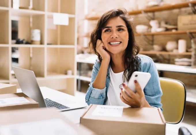 Smiling small business owner holding a smartphone at a desk with a laptop and shipping boxes in a shop