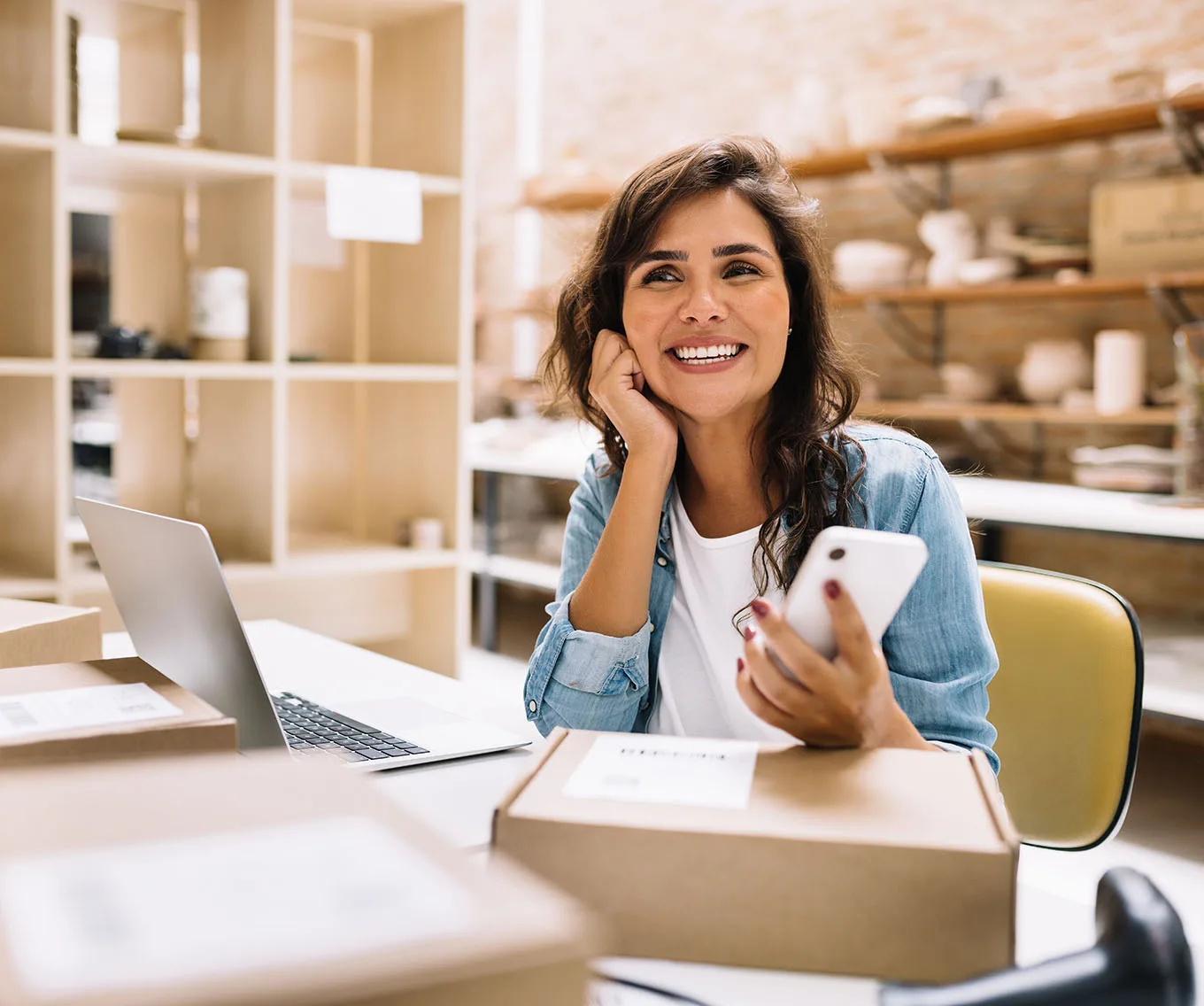 Smiling small business owner holding a smartphone at a desk with a laptop and shipping boxes in a shop