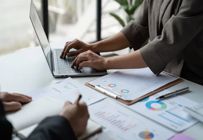 Two professionals collaborating at a desk with a laptop, charts, and financial report documents