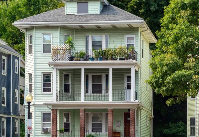 Three-story light green investment property with stacked balconies, front porches and a pitched roof, surrounded by trees.