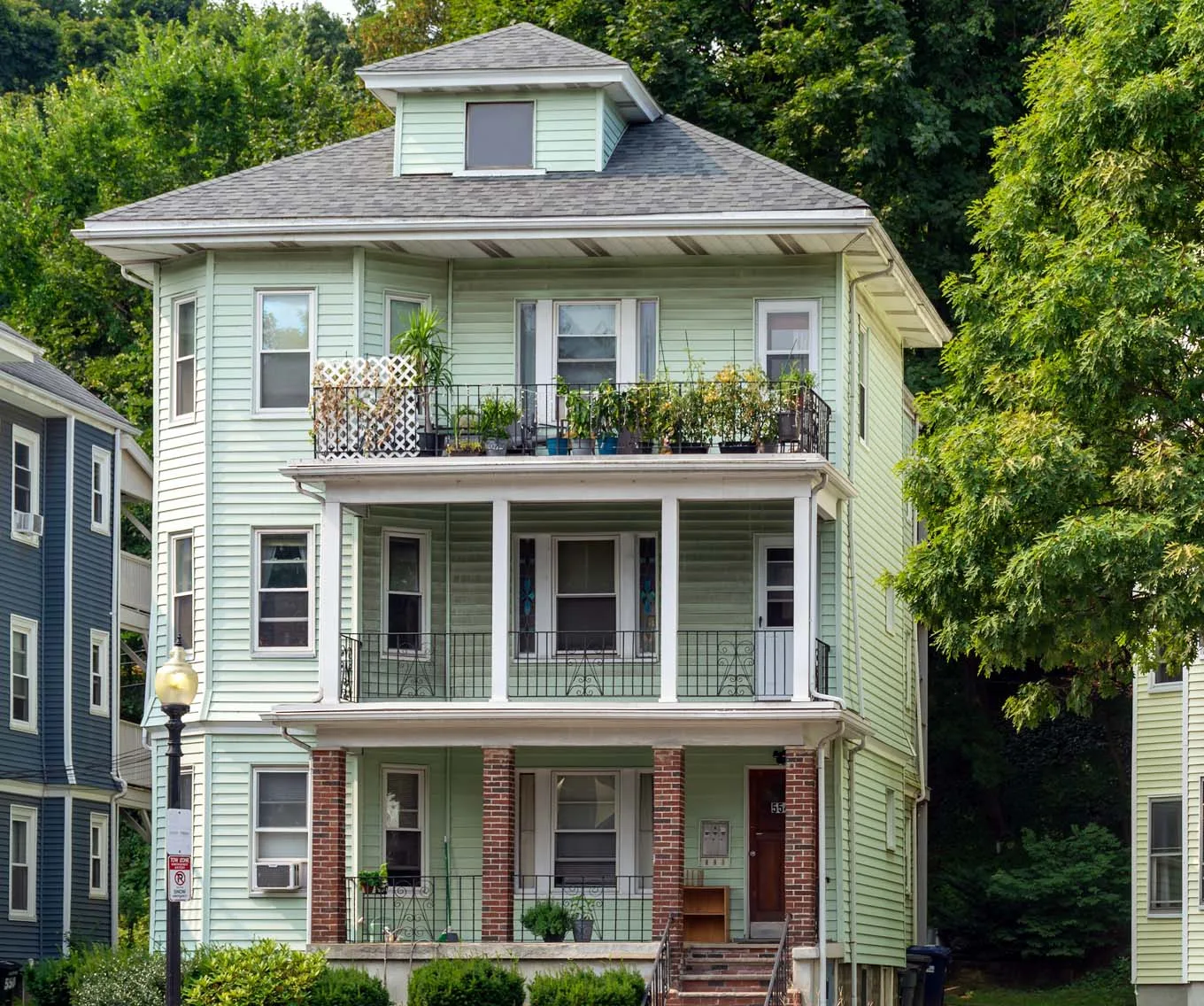 Three-story light green investment property with stacked balconies, front porches and a pitched roof, surrounded by trees.