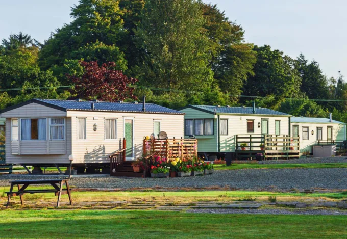 Three manufactured homes with porches beside a gravel lane, with grass and trees surrounding the community.