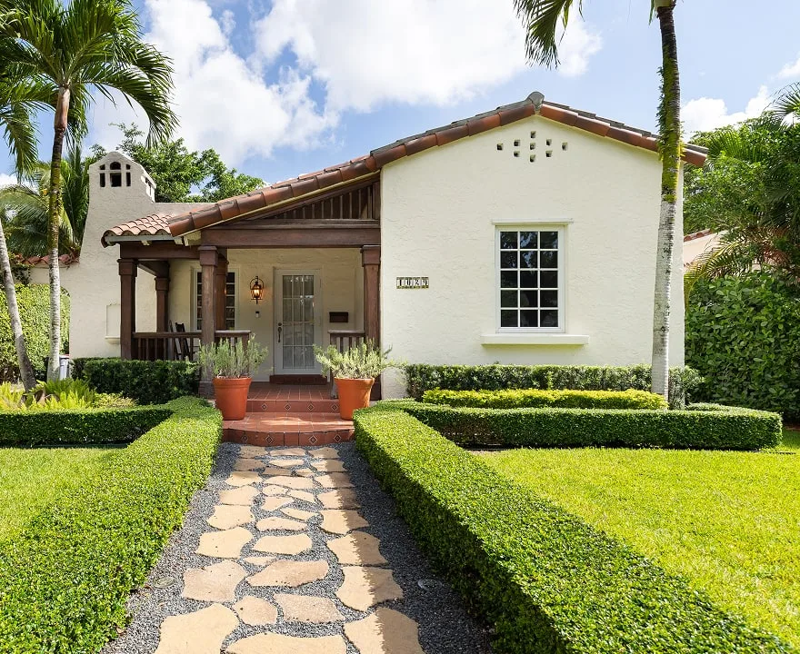 White stucco Florida home with a red tile roof, covered front porch, palm trees and a stone walkway bordered by manicured hedges.