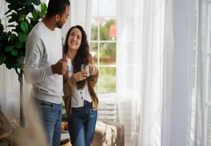 Couple smiling and talking together at home by a sunny window, each holding a coffee mug
