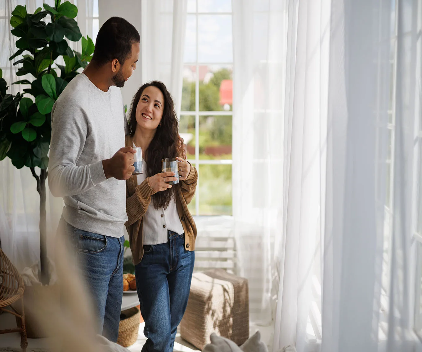Couple smiling and talking together at home by a sunny window, each holding a coffee mug