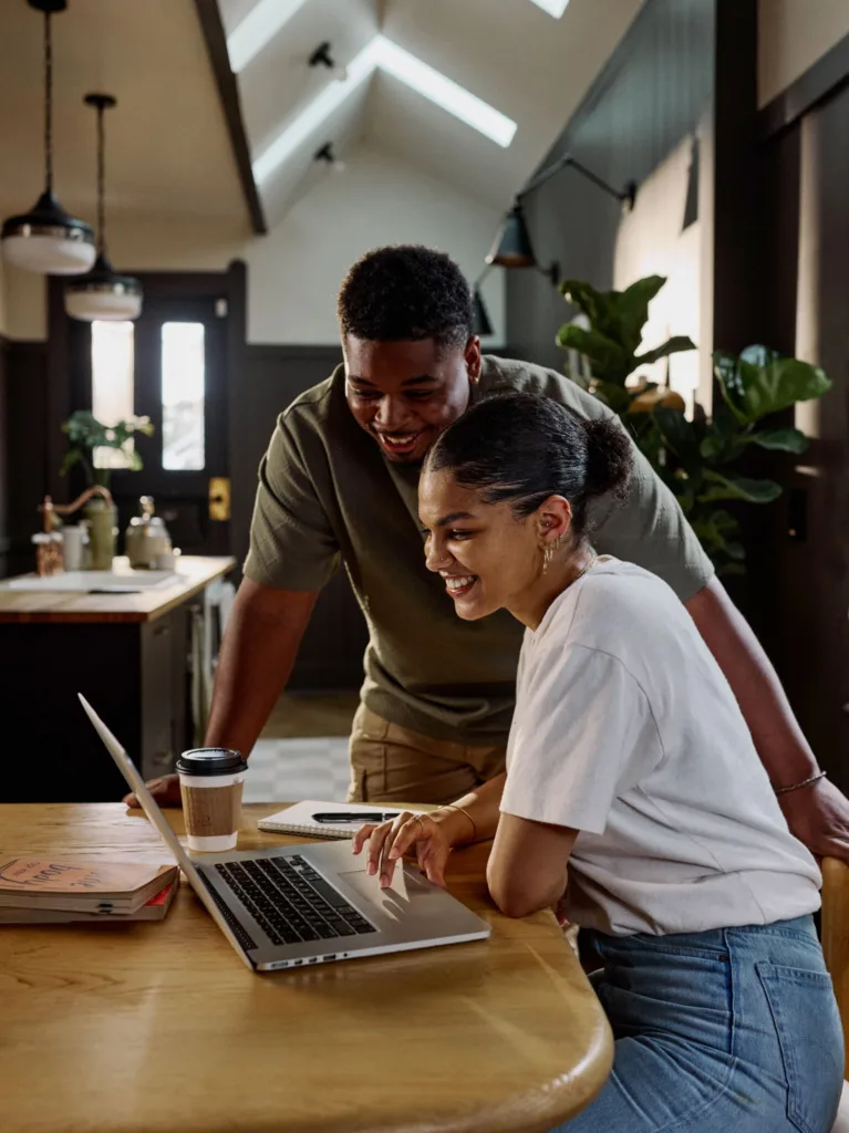 Two people smiling and looking at a laptop together at a kitchen table, with coffee and notebooks nearby