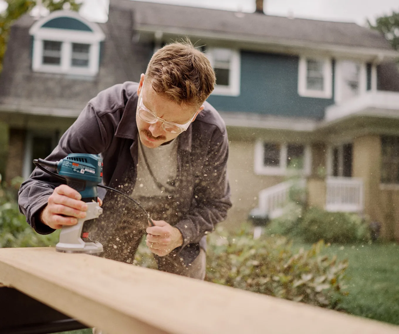 Person using an electric sander to smooth a wooden board outdoors in front of a house, illustrating sweat equity through DIY home improvements.
