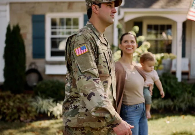 Service member in camouflage uniform standing outside a suburban home with a woman holding a young child.