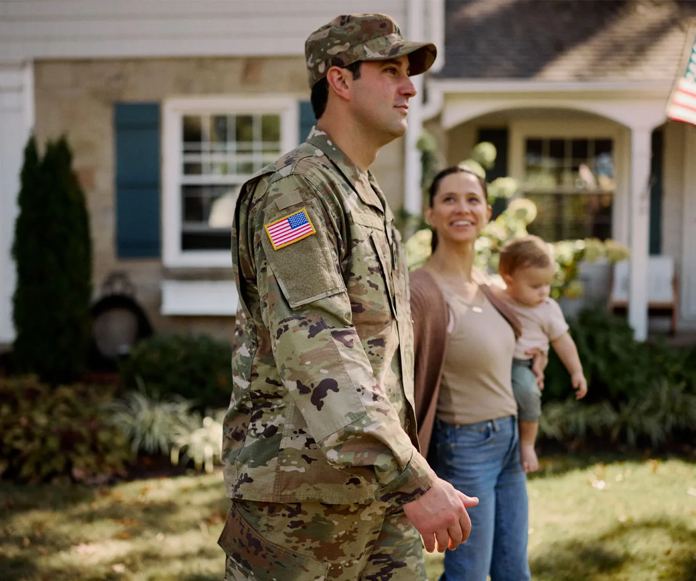 Service member in camouflage uniform standing outside a suburban home with a woman holding a young child.