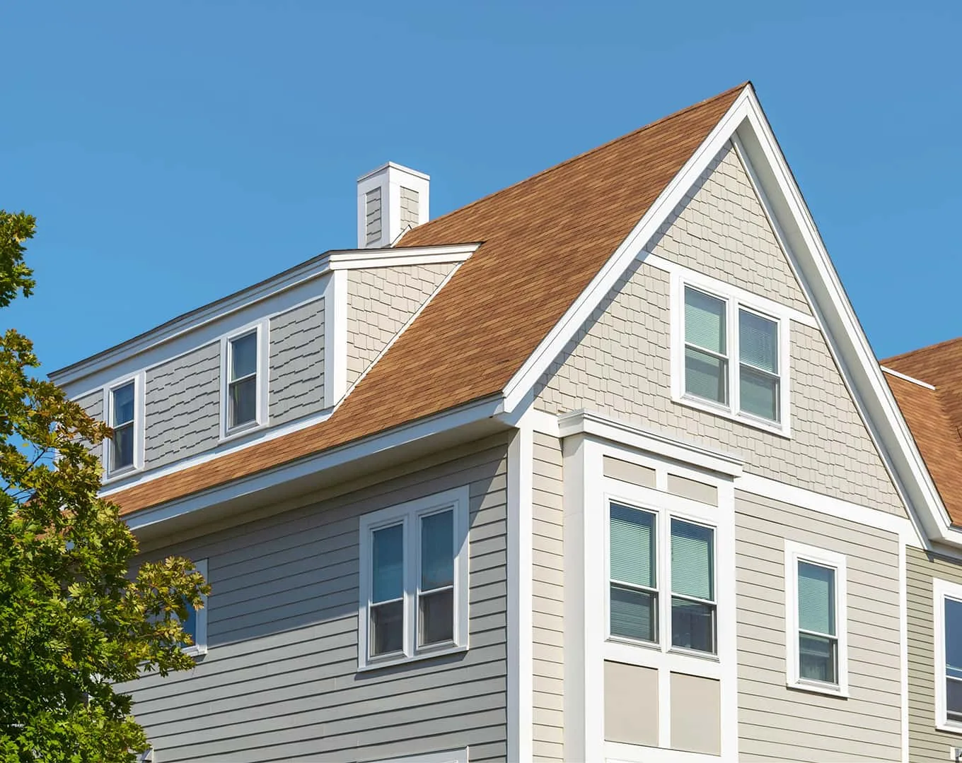 Upper exterior of a gray house with white trim, a brown shingle roof and windows beneath a clear blue sky.