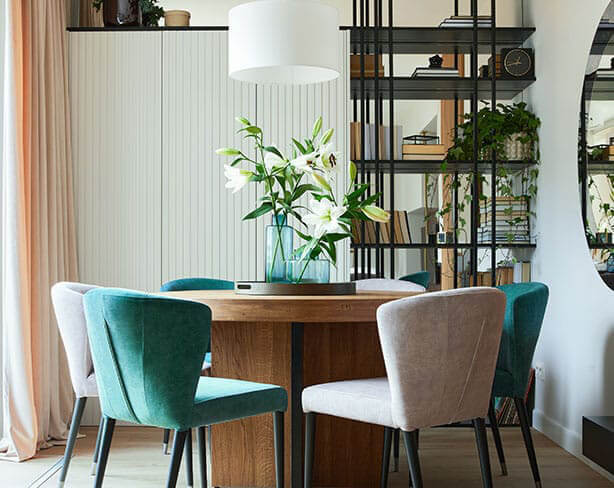 Interior of a home. A round dining room table in the forefront of the image with chairs around it. A bookshelf in the background to the right and storage furniture to the back left.